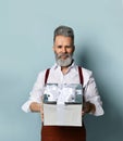 Gray-haired man in white shirt, brown pants and suspenders. Smiling, holding silver gift box, posing against blue background Royalty Free Stock Photo