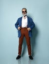 Senior man in white shirt, jacket, brown pants, black loafers. Straightening sunglasses, hand in pocket, posing on blue background