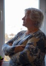A gray-haired elderly woman stands at the window with her arms crossed over her chest. Royalty Free Stock Photo