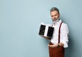 Gray-haired man in white shirt, brown pants and suspenders. Smiling, holding silver gift box, posing against blue background Royalty Free Stock Photo