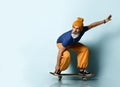 Aged man in t-shirt, orange pants, hat, gumshoes. Riding black skateboard, looking at it, posing on blue background Royalty Free Stock Photo