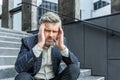 A gray-haired senior male businessman sits outside an office center on the stairs. Upset, holding his head. Tired of Royalty Free Stock Photo