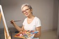 Gray-haired sophisticated lady in glasses stands in front of easel