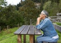 A gray-haired woman sits at a wooden table in the countryside at the edge of the forest with a kind look.