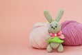 Gray knitted hare stands on the background of multi-colored balls of yarn on a pink background. There is a place for text