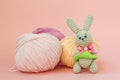 Gray knitted hare stands on the background of multi-colored balls of yarn on a pink background. There is a place for text