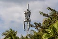 Gray metal cell tower carrying antennas of cellular networks on the blue sky with white clouds background with green trees and