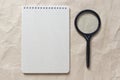 Gray notepad with white coiled spring and magnifieron a background of beige crumpled craft paper