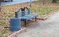 A gray painted wooden bench and a trash can in an autumn park near the footpath Royalty Free Stock Photo