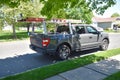 Gray pickup truck with ladders on a rack and equipment in the bed parked by a road curb