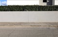 Gray plaster fence of modern house with railing and hedge on top. Wall and sky on behind. Concrete sidewalk and road in front