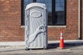Gray plastic portable bathroom sitting in parking lot in front of brick building - female symbol on door and contruction cone next
