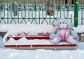 A gray plush toy hare with a pink scarf sits on a bench in the snow in winter, forgotten by a child. The theme of sadness, loneli