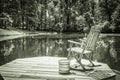 Gray scale shot of a wooden chair on a wooden dock in the middle of a lake surrounded by trees Royalty Free Stock Photo