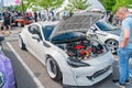A gray sports car sits on pavement, its hood open to reveal engine components, amidst a busy outdoor setting with
