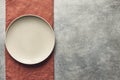 Gray textured concrete background for design with beige empty food plate on terracotta linen napkin. Copy space