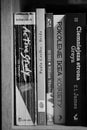 Grayscale closeup view of a row of books arranged on a wooden shelf.