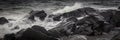 Grayscale panoramic shot of the ocean waves reaching the stones on the shore