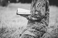 Grayscale shot of a young soldier kneeling while holding an open bible in a field