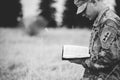 Grayscale shot of a young soldier reading a bible in a field