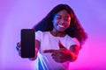 Great mobile offer. Happy young black lady pointing finger at smartphone, advertising new app in neon light Royalty Free Stock Photo