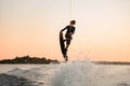 Great view of active male rider holds rope and masterfully jumping over splashing wave on wakeboard at sunset.