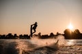 Great view of dark silhouette of active male rider holds rope and making extreme jump on wakeboard at sunset.