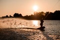 Great view of dark silhouette of active male rider holds rope riding on wakeboard on water surface at sunset.