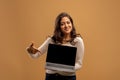 Great website. Cheerful arab lady pointing at laptop with blank screen and smiling, brown background, mockup, free space