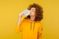 Greed for money. Portrait of avaricious curly-haired woman in urban style hoodie smelling dollar banknotes Royalty Free Stock Photo