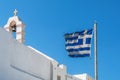 Greek flag waving on a must under strong wind against blue sky background, before a white Orthodox church, with belltower and Royalty Free Stock Photo