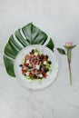 Greek salad with Pink peony flower and monstera leaf under plate, marble background