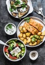 Greek style lunch table - baked lemon salmon with potatoes, greek salad, grilled eggplant with tzadziki sauce on dark background,