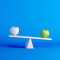 Green apple sitting on seesaw with white apple on opposite end on blue background