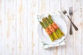 Baked bundles of green asparagus wrapped in bacon in a ceramic plate on a white wooden table, top view Royalty Free Stock Photo