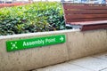 Green assembly point sign with people icon and arrow on wall beside wooden bench at shopping centre. Royalty Free Stock Photo