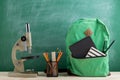 green backpack, microscope, black notebooks and pencils on the background of the blackboard