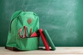 green backpack, red scissors and notebook and book on the background of the blackboard