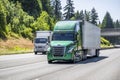 Green big rig semi truck with dry van semi trailer and small rig cab over semi truck with box trailer running side by side on the Royalty Free Stock Photo