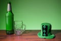 Green bottle of lager and glass and green hat with shamrock on a wooden bar surface. Green background. Saint Patrick theme