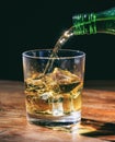 Green bottlepouring whiskey in crystal glass with ice cubes on wooden table Royalty Free Stock Photo