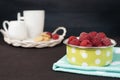 Green bowl with polka dot full with raspberries. Cup of coffee, cookies a jug of milk on a straw tray. Black background Royalty Free Stock Photo