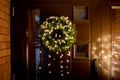 Green color spruce tree tips Christmas Wreath with warm lights and white felt pom-poms on home entryway.