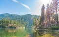 Green colored lake surrounded by pine forest with bright sunshine and blue sky