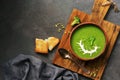 Green cream broccoli soup with sour cream and pumpkin seeds in a ceramic bowl on a cutting board, dark rustic background.