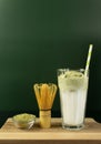 Green dalgona in a tall glass with paper straw, matcha whisk and matcha tea powder on a wooden board with a copy space