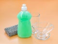 Green dish washing liquid in a transparent plastic bottle, silver foam sponge and glassware on a pastel orange background. Kitchen