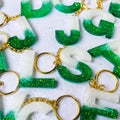 green glitter letters with beads. Golden keychain on white paper background with dried flowers. Top view. Resin craft