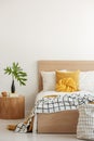 Green leaf in vase on wooden nightstand next to Yellow pillows and blanket on white single wooden bed