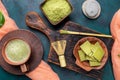 Green matcha tea and powder in brown ceramic cups and matcha chocolate on wooden serving board on emerald background.